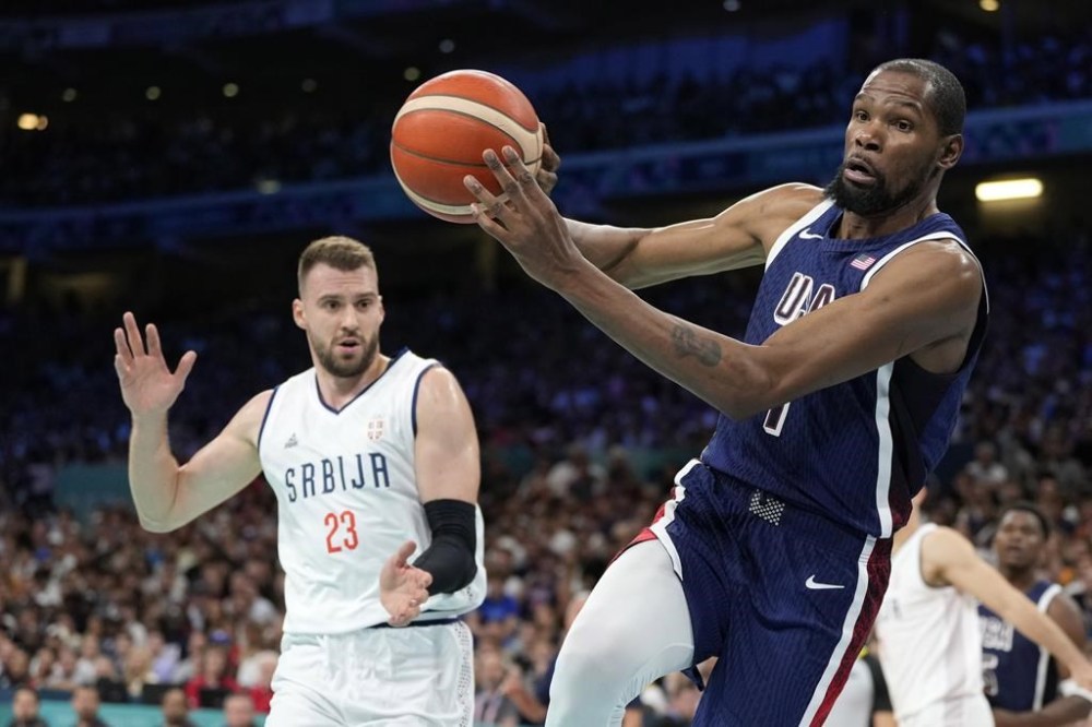 Kevin Durant, right, of the United States, passes the ball as he heads out of bounds while under pressure from Marko Guduric, of Serbia, in a men's basketball game at the 2024 Summer Olympics, Sunday, July 28, 2024, in Villeneuve-d'Ascq, France. (AP Photo/Michael Conroy)