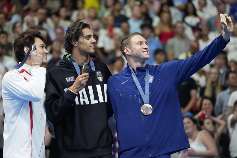 Gold medalist Thomas Ceccon, of Italy, stands on the podium with silver medalist Xu Jiayu, left, of China, and bronze medalist Ryan Murphy, right, of the United States, for a selfie following the men's 100-meter backstroke final at the 2024 Summer Olympics, Monday, July 29, 2024, in Nanterre, France. (AP Photo/Matthias Schrader)