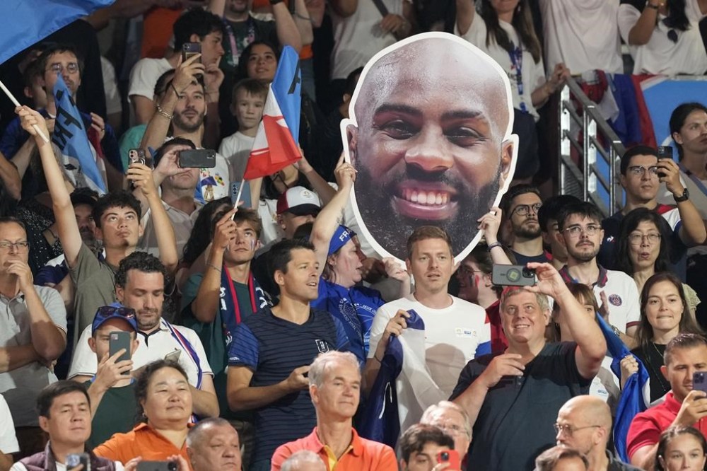 Fans of France cheer holding banner depicts France's Teddy Riner during men's +100 kg elimination round match in the team judo competition, at Champ-de-Mars Arena, during the 2024 Summer Olympics, Friday, Aug. 2, 2024, in Paris, France. (AP Photo/Eugene Hoshiko)