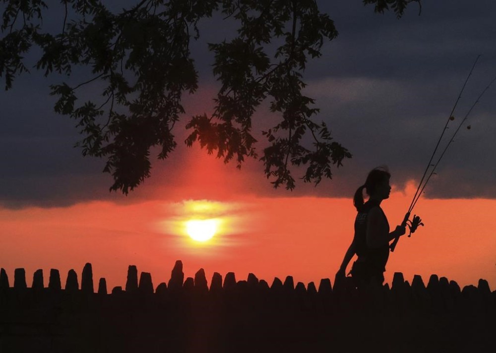 A person walks with fishing gear as the sun is obscured from the wildfires in the western United States and Canada in Oconomowoc, Wis. Saturday, Aug. 3, 2024. (John Hart/Wisconsin State Journal via AP)