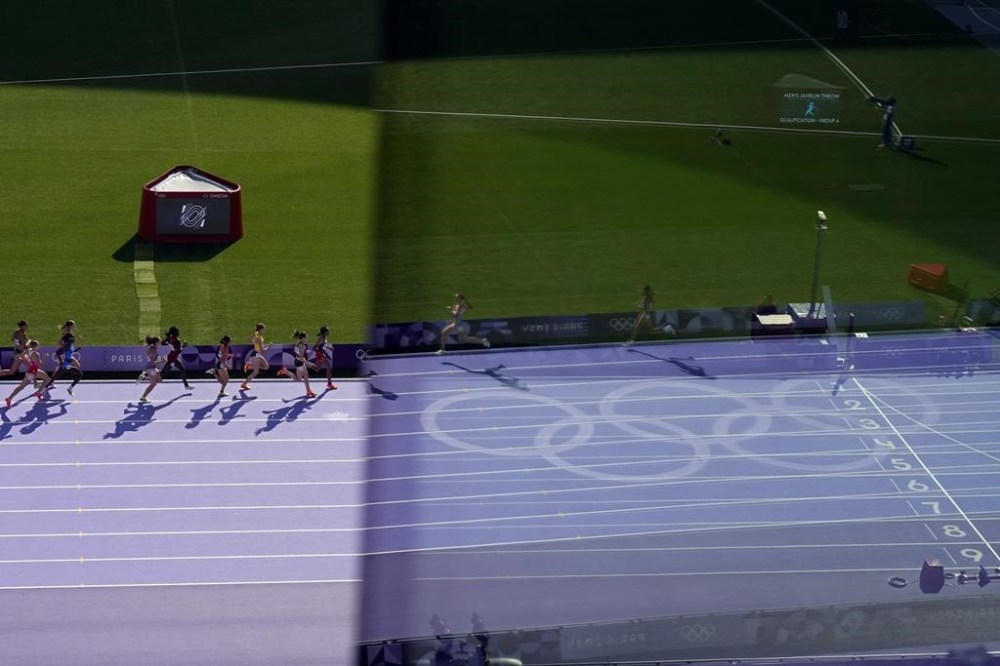 Athletes run in a women's 1500 meters round 1 heat, while the finish line is seen through a window glass, at the 2024 Summer Olympics, Tuesday, Aug. 6, 2024, in Saint-Denis, France. (AP Photo/Ashley Landis)