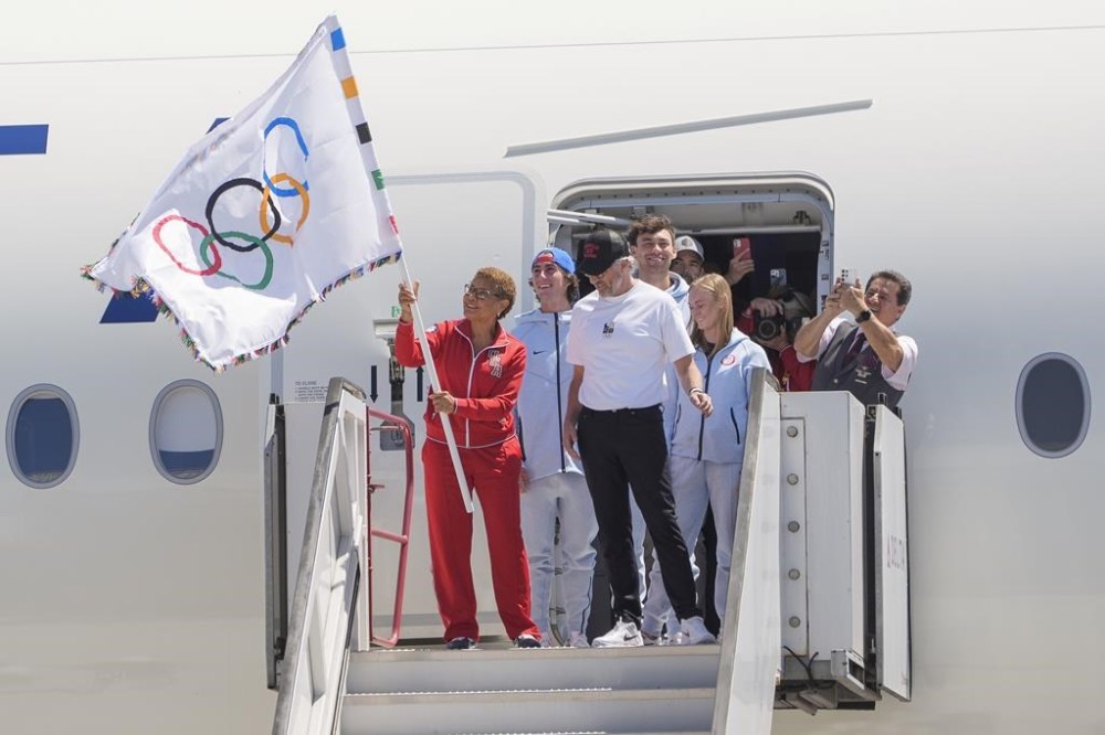 Los Angeles Mayor Karen Bass holds up the Olympic flag, with LA28 chairman Casey Wasserman, right, and members of Team USA athletes at Los Angeles International Airport on Monday, Aug. 12, 2024. (AP Photo/Damian Dovarganes)