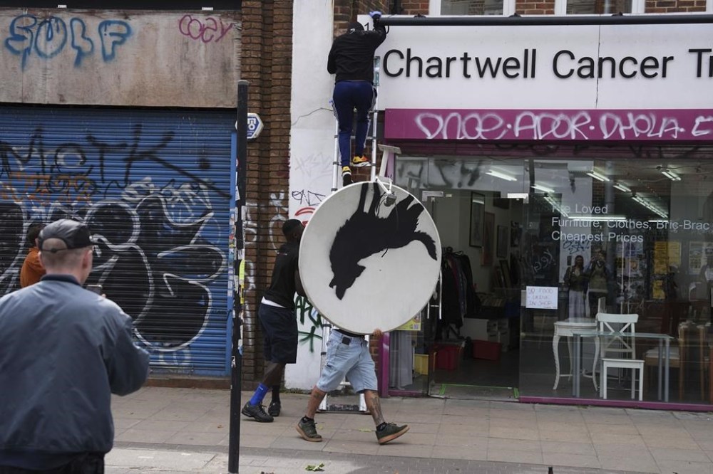 People remove a new artwork by Banksy, depicting a howling wolf painted on a satellite dish that was placed on a shop roof in Peckham, south London, Thursday Aug. 8, 2024. (Jordan Pettitt/PA via AP)