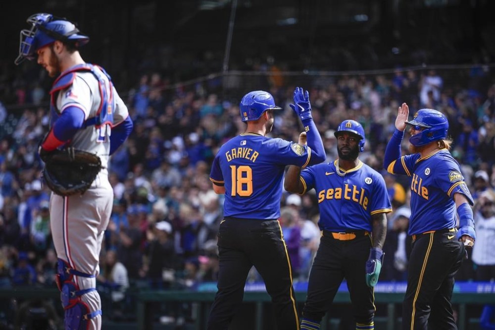Seattle Mariners' Mitch Garver (18) greets teammates Randy Arozarena, center, and Justin Turner, right, who both scored on his three-run home run, as Texas Rangers catcher Jonah Heim, left, looks down during the first inning of a baseball game Sunday, Sept. 15, 2024, in Seattle. (AP Photo/Lindsey Wasson)