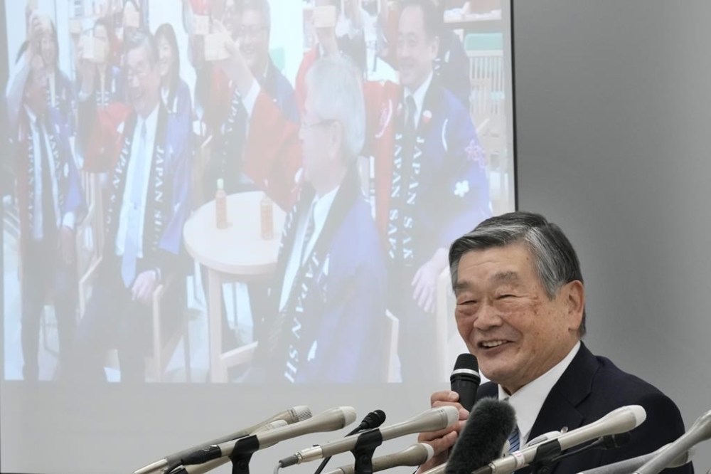 Shinuemon Konishi, president of the Preservation Society of Japanese Koji-based Sake-making craftsmanship, responds to a reporter's question during a news conference in Tokyo, Thursday, Dec. 5, 2024, as a video feed, background, shows a group of Japanese people including Konishi early in the morning celebrate UNESCO's recognition of Japan's sake making as