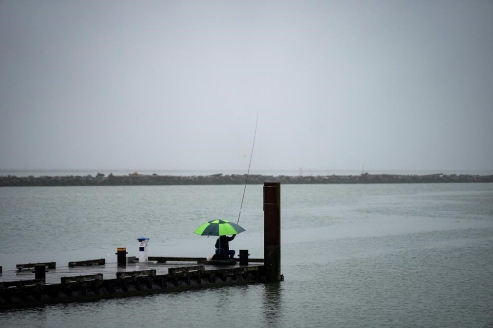 A man sits under an umbrella on a dock while fishing for perch and whitefish on the Fraser River as rain falls, in Richmond, B.C., Monday, July 24, 2023. THE CANADIAN PRESS/Darryl Dyck