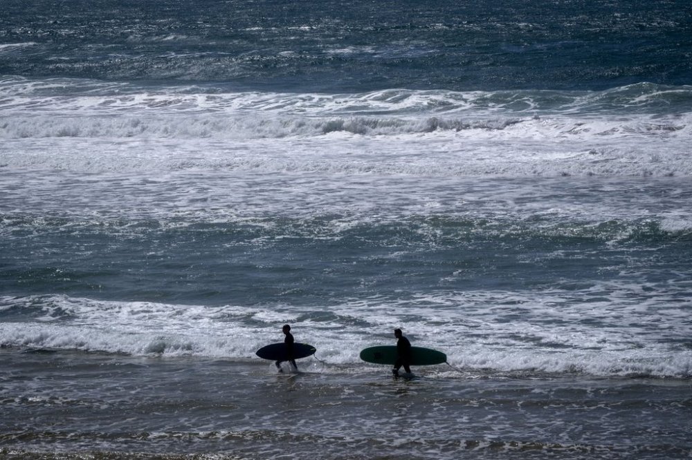 FILE - Two surfers walk with their boards on the beach of the Atlantic Ocean in Sines, Portugal, April 1, 2024. (AP Photo/Michael Probst, File)