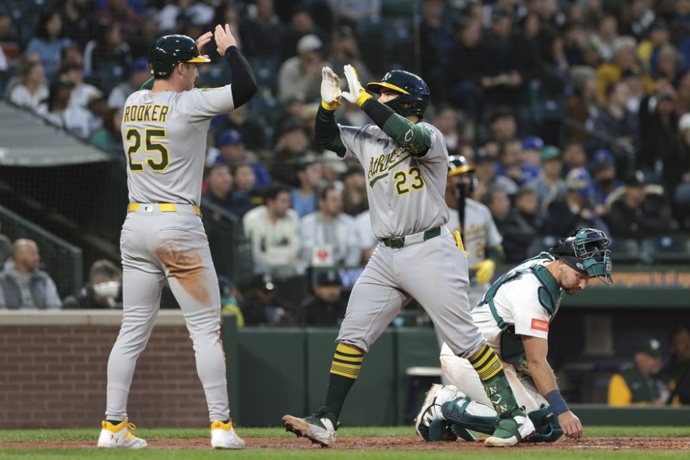 Athletics' Brent Rooker (25) celebrates with Shea Langeliers (23) at home plate after they scored on Langeliers' two-run home run as Seattle Mariners catcher Cal Raleigh, right, looks on during the fourth inning of a baseball game, Saturday, March 29, 2025, in Seattle. (AP Photo/Jason Redmond)