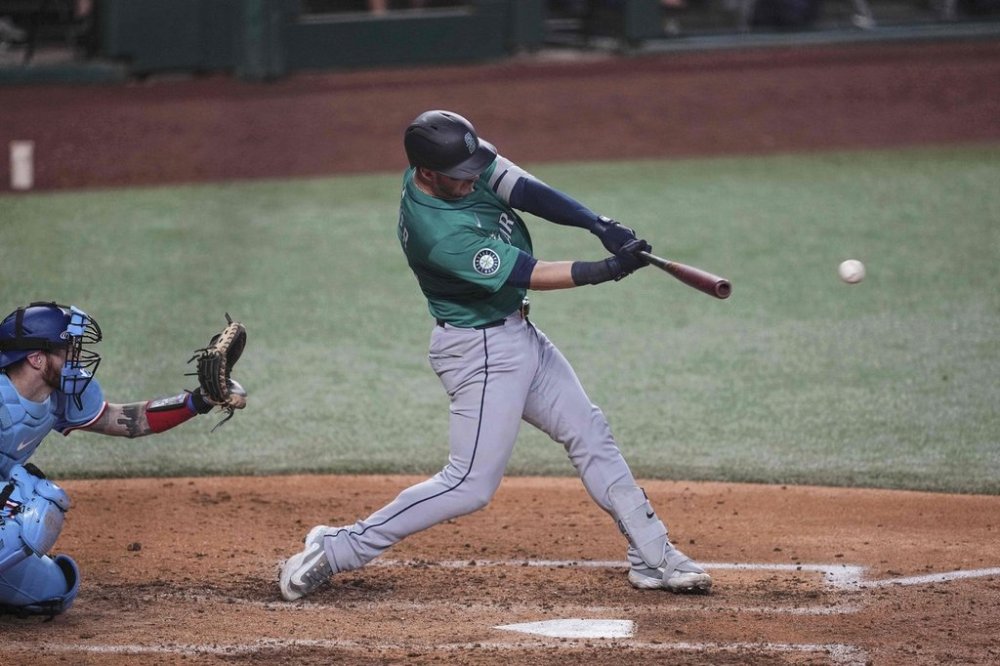 Seattle Mariners' Mitch Garver follows through on an RBI single that scored Randy Arozarena as Texas Rangers catcher Jonah Heim, left, looks on in the sixth inning of a baseball game Sunday, June 29, 2025, in Arlington, Texas. (AP Photo/Tony Gutierrez)