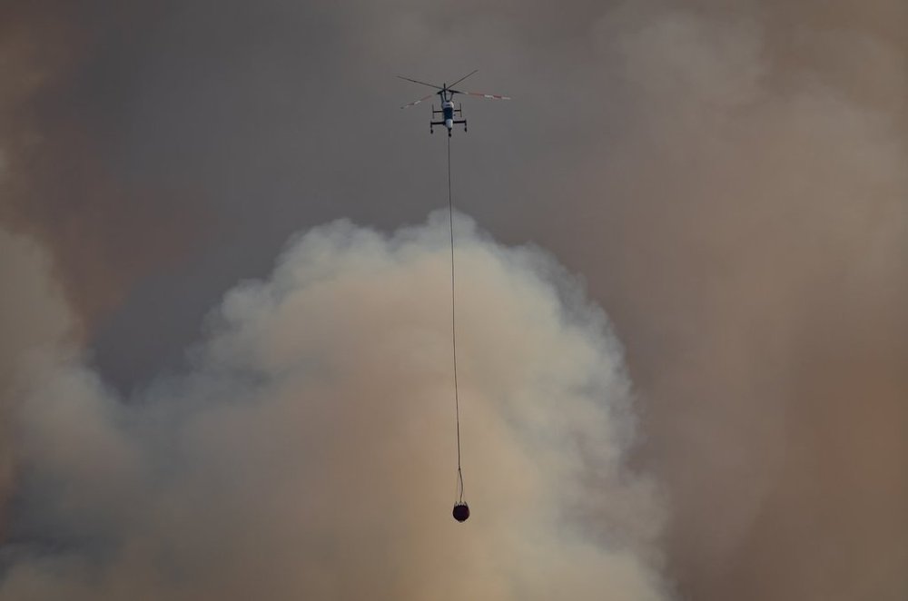 A helicopter with a water bucket flies past smoke rising into the air while battling the July Mountain wildfire along the Coquihalla Highway south of Merritt, B.C., on August 11, 2021. THE CANADIAN PRESS/Darryl Dyck