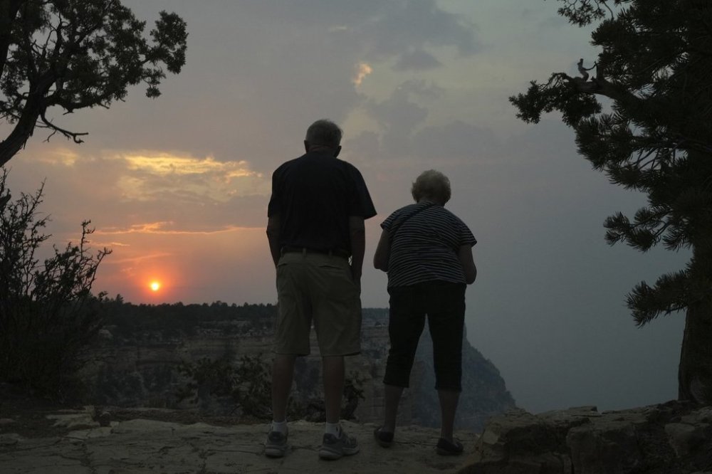 The setting sun peeks through the smoke from the Dragon Bravo Fire as it moves over the Grand Canyon from the North Rim as two tourists take in the scene from a South Rim overlook Monday, July 14, 2025, in Grand Canyon National Park, Ariz. (AP Photo/Ross D. Franklin)
