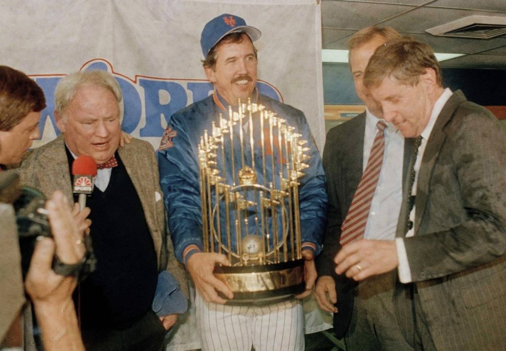 FILE - New York Mets Manager Davey Johnson, center, holds baseball's World Series trophy on the podium after the Mets defeated the Boston Red Sox in Game 7, at Shea Stadium in New York, Oct. 27, 1986. Mets General Manager Frank Cashen, left, Baseball Commissioner Peter Ueberroth, partially hidden, and Mets President Fred Wilpon, right, join the celebration in the locker room the clubhouse. (AP Photo/Ray Stubblebine, File)