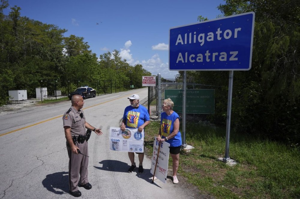 A police officer urges Art Sennholtz, 80, center, and Christy Howard, 70, of Just Us Volusia to be careful of fast-moving traffic as they hold protest signs outside the entrance to an immigration detention center in the Florida Everglades known as