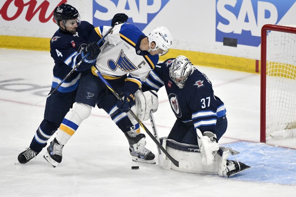 Jets goaltender Connor Hellebuyck (37) makes a save on St. Louis Blues captain Brayden Schenn (10) as Neal Pionk (4) defends during first period of an NHL playoff game in Winnipeg on Monday, April 21, 2025. THE CANADIAN PRESS/Fred Greenslade