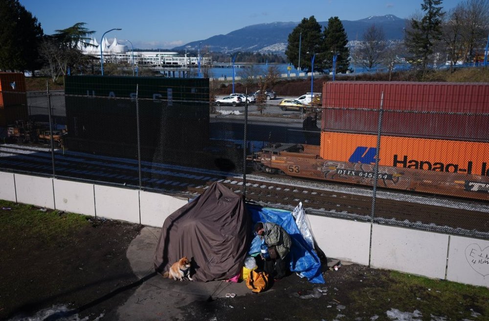 A man living in a tent prepares to give his dog water as shipping containers being moved at port sit on railroad cars, in Vancouver, on Friday, Feb. 14, 2025. THE CANADIAN PRESS/Darryl Dyck