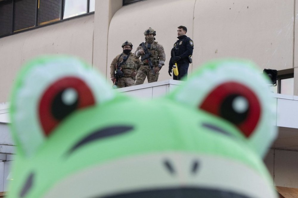 Law enforcement officers watch from a ledge of a United States Immigration and Customs Enforcement (ICE) facility as a protester stands outside in an inflatable frog costume on Tuesday, Oct. 21, 2025, in Portland, Ore. (AP Photo/Jenny Kane)