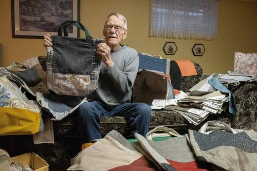 SVJETLANA MLINAREVIC WINNIPEG FREE PRESS Retired gym teacher Peter Dick sits among the fabric scraps and samples holding a shopping bag he made from those pieces of fabric, which are used to raise money for South East Helping Hands in Stienbach on Monday, Dec. 22, 2025. His shopping bags sell for $5 each and are made from the fabric scraps and fabric samples from Dufresne Furniture and Appliances stores in Steinbach and Thunder Bay. He has been sewing the shopping bags for more than 18 years and has sewn 850 bags to date. After retirement, he began sewing quilts and then later switched to the shopping bags. In total, he has been sewing for more than 25 years.