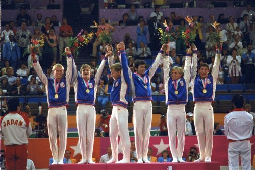 FILE - Members of the U.S. gymnastics team, from left: Bart Conner, Peter Vidmar, Jim Hartung, Mitch Gaylord, Scott Johnson and Tim Daggett, celebrate their gold medals as they stand on the winners' platform July 31, 1984 after they defeated world champion China to win the first U.S. gold medal in gymnastics in 80 years. (AP Photo/Lionel Cironneau, file)