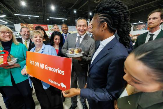 On Wednesday afternoon, Premier Wab Kinew, surrounded by most of his cabinet at the Red River Co-op on Grant Avenue, prepares to take a bite out of a rotisserie chicken to celebrate removing the PST from groceries. (Mike Deal / Free Press)