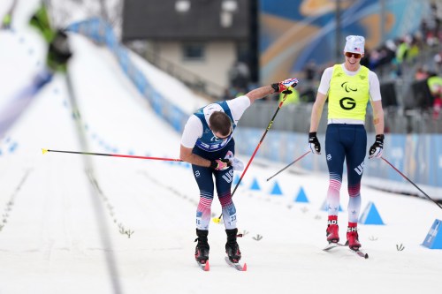 Jake Adicoff, of the United States, crosses the finish line, as his guide Reid Goble at right looks on, to win the gold medal in the cross country skiing men's 10Km interval start classic vision impaired final at the 2026 Winter Paralympics, in Tesero, Italy, Wednesday, March 11, 2026. (AP Photo/Evgeniy Maloletka)