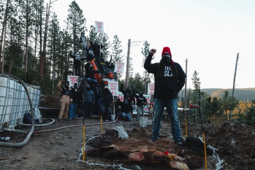 A masked demonstrator stands with their fist in the air, wearing a Land Back beanie in front of signs reading Protect Pe'Sla Now on April 30, 2026, in Pennington County, S.D. (Angel White Eyes via AP)