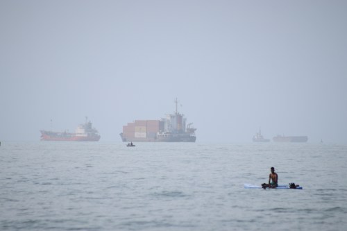 FILE - A man sits on a surfboard as a container ship and other commercial vessels appear anchored in the Strait of Hormuz off Bandar Abbas, Iran, Monday, July 27, 2026. (Razieh Poudat/ISNA via AP, File)